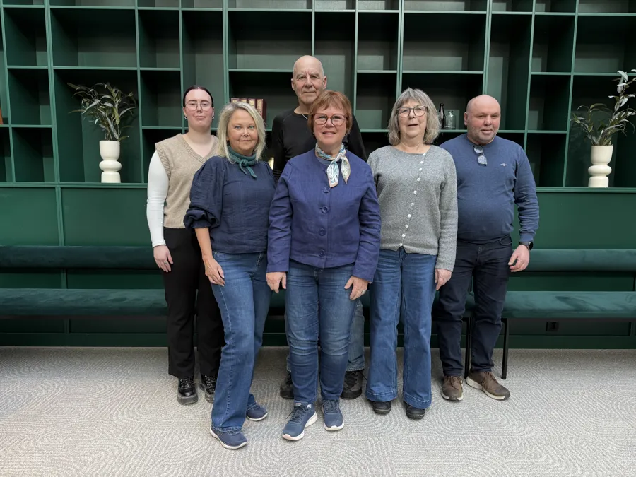 En gruppe på seks personer står samlet innendørs foran en stor, mørkegrønn hyllevegg som dekker hele bakgrunnen. Hyllene inneholder enkelte dekorative elementer, blant annet hvite vaser med planter og noen få bøker. Personene står i to rader, med fire foran og to bak. De er kledd i hverdagslige klær i blå og grå toner, inkludert jeans, strikkegensere, skjorter og skjerf. Gulvet under dem er lyst og mønstret, og bak gruppen står en lang grønn benk integrert i veggen. Lyset i rommet er mykt og jevnt, og helhetsinntrykket er et uformelt gruppebilde i et moderne, stilrent interiør.