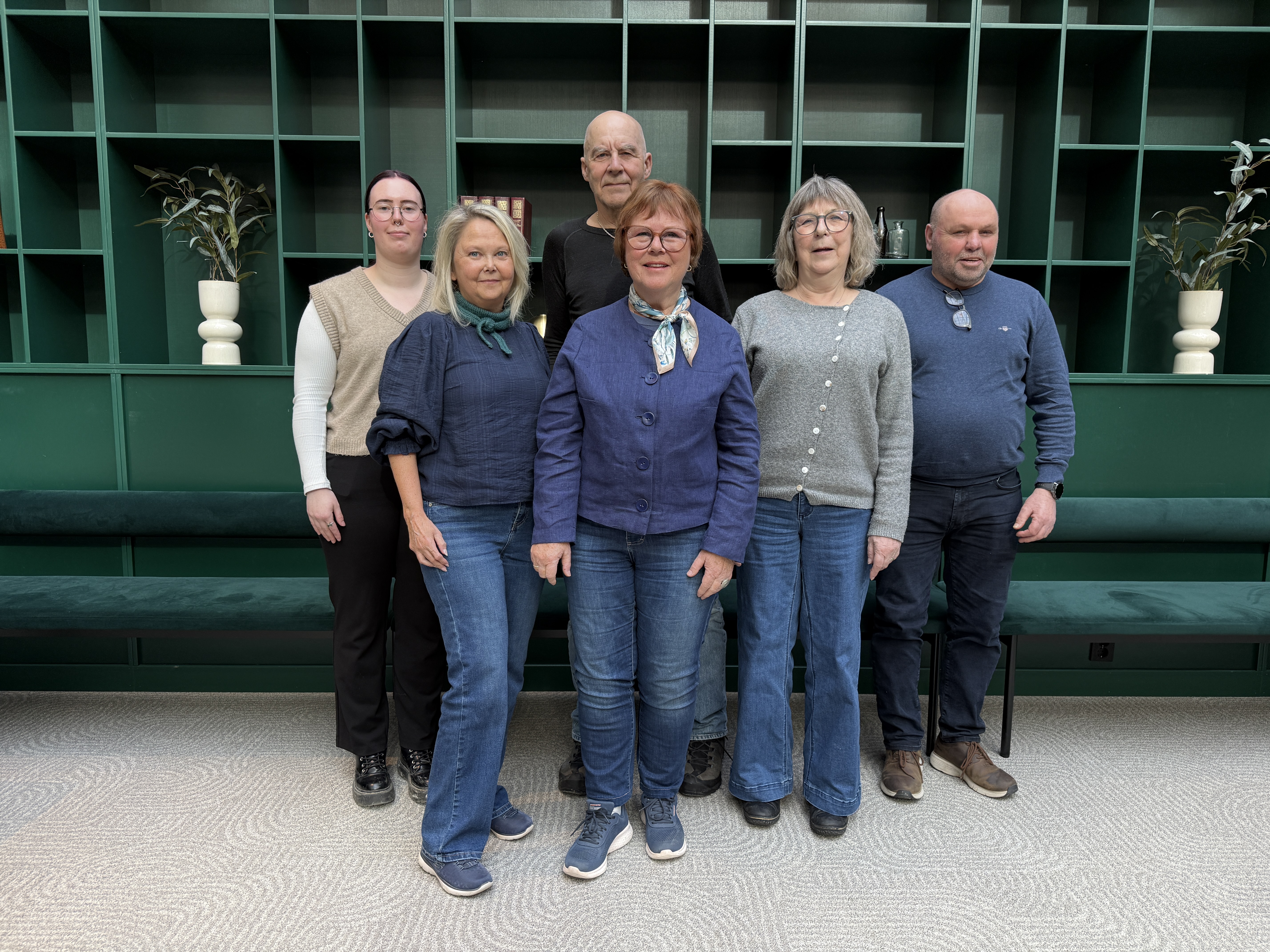 En gruppe på seks personer står samlet innendørs foran en stor, mørkegrønn hyllevegg som dekker hele bakgrunnen. Hyllene inneholder enkelte dekorative elementer, blant annet hvite vaser med planter og noen få bøker. Personene står i to rader, med fire foran og to bak. De er kledd i hverdagslige klær i blå og grå toner, inkludert jeans, strikkegensere, skjorter og skjerf. Gulvet under dem er lyst og mønstret, og bak gruppen står en lang grønn benk integrert i veggen. Lyset i rommet er mykt og jevnt, og helhetsinntrykket er et uformelt gruppebilde i et moderne, stilrent interiør.