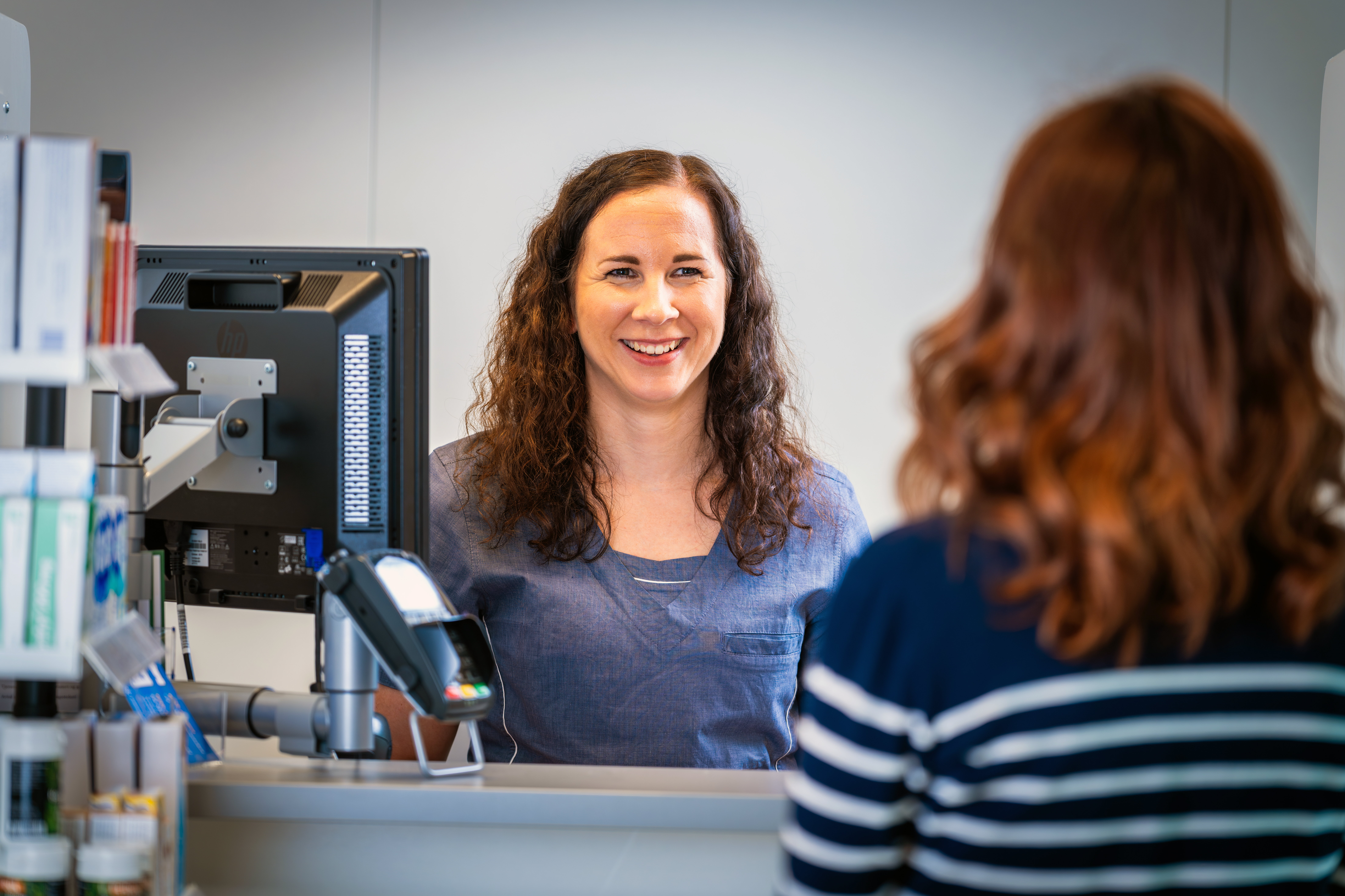 Person bak en apotekdisk snakker med en kunde som står på motsatt side. En dataskjerm, betalingsterminal og legemidler er synlige på disken. Bildet er tatt innendørs i et apotekmiljø.