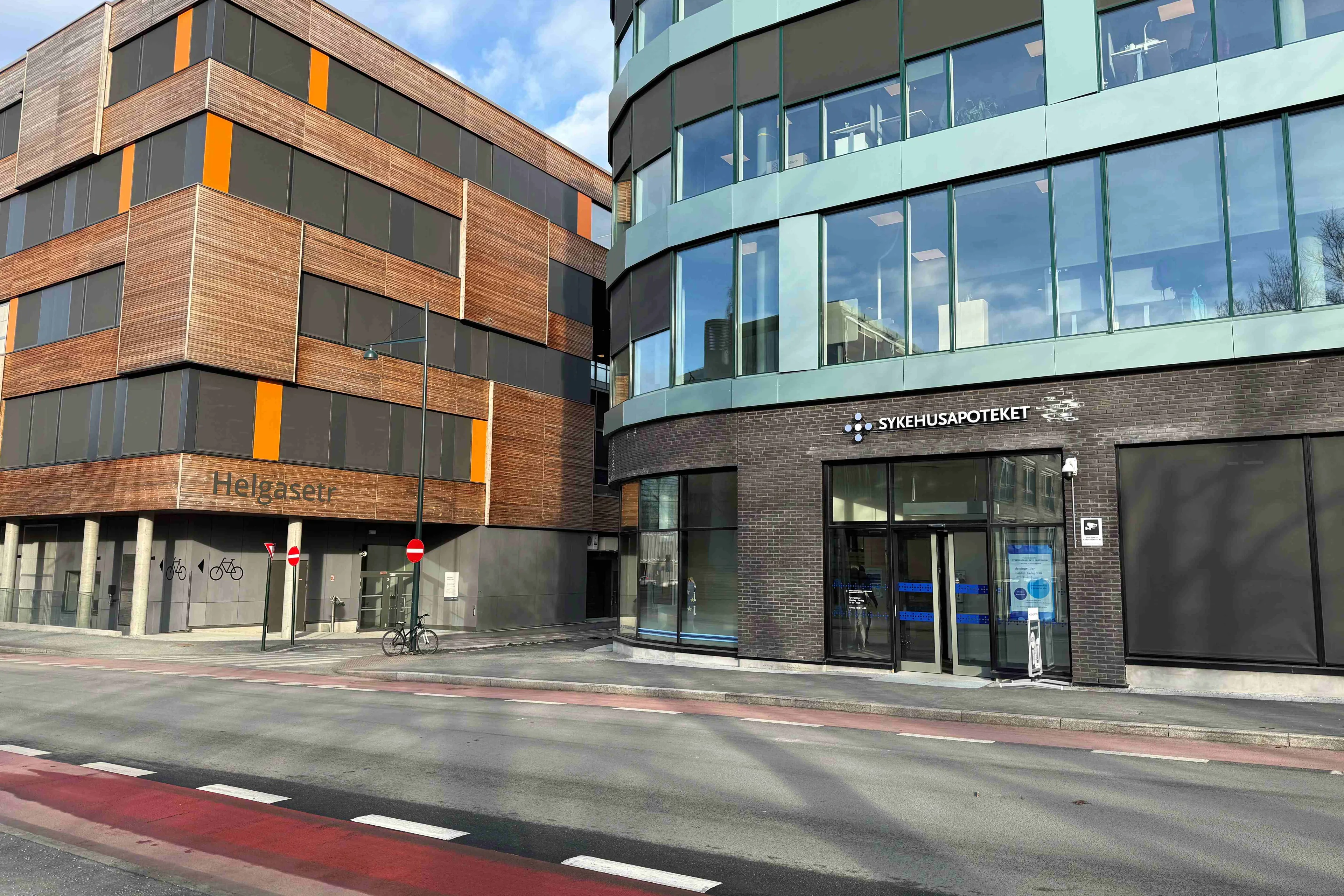 Bildet viser to moderne bygninger ved en gate. Til høyre står en grønn bygning med store glassvinduer, hvor inngangen er merket med teksten «SYKEHUSAPOTEKET» over døren. Dørene er i glass, og det er mulig å se inn i lokalet. Til venstre står en mer kantete bygning med brun fasade i tre og flere vertikale felt i oransje. På veggen står navnet «Helgasetr». I forgrunnen går en vei med fortau langs bygningene.