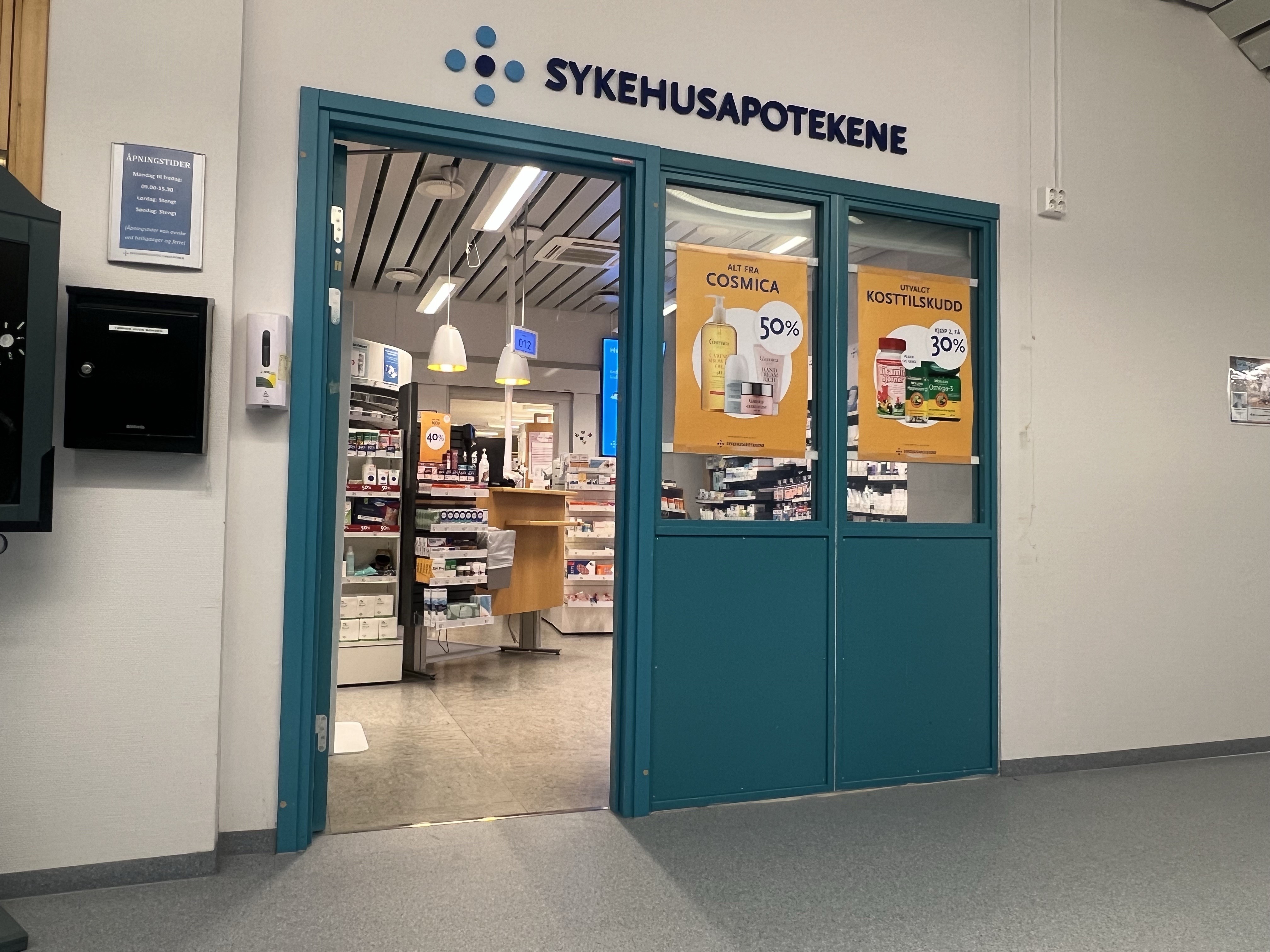 Inngang til Sykehusapotekene med turkis dør- og vindusramme. Over inngangen står logoen og teksten «SYKEHUSAPOTEKENE». Gjennom de store glassfeltene er deler av apoteklokalet synlige, inkludert betjeningsdisk, varehyller og belysning. På vinduene henger kampanjeplakater for hudpleieprodukter og kosttilskudd med rabattinformasjon. Til venstre for inngangen er det oppslag om åpningstider og en dispenser montert på veggen. Lokalet ligger i en sykehuskorridor med lyse vegger og grått gulv.
