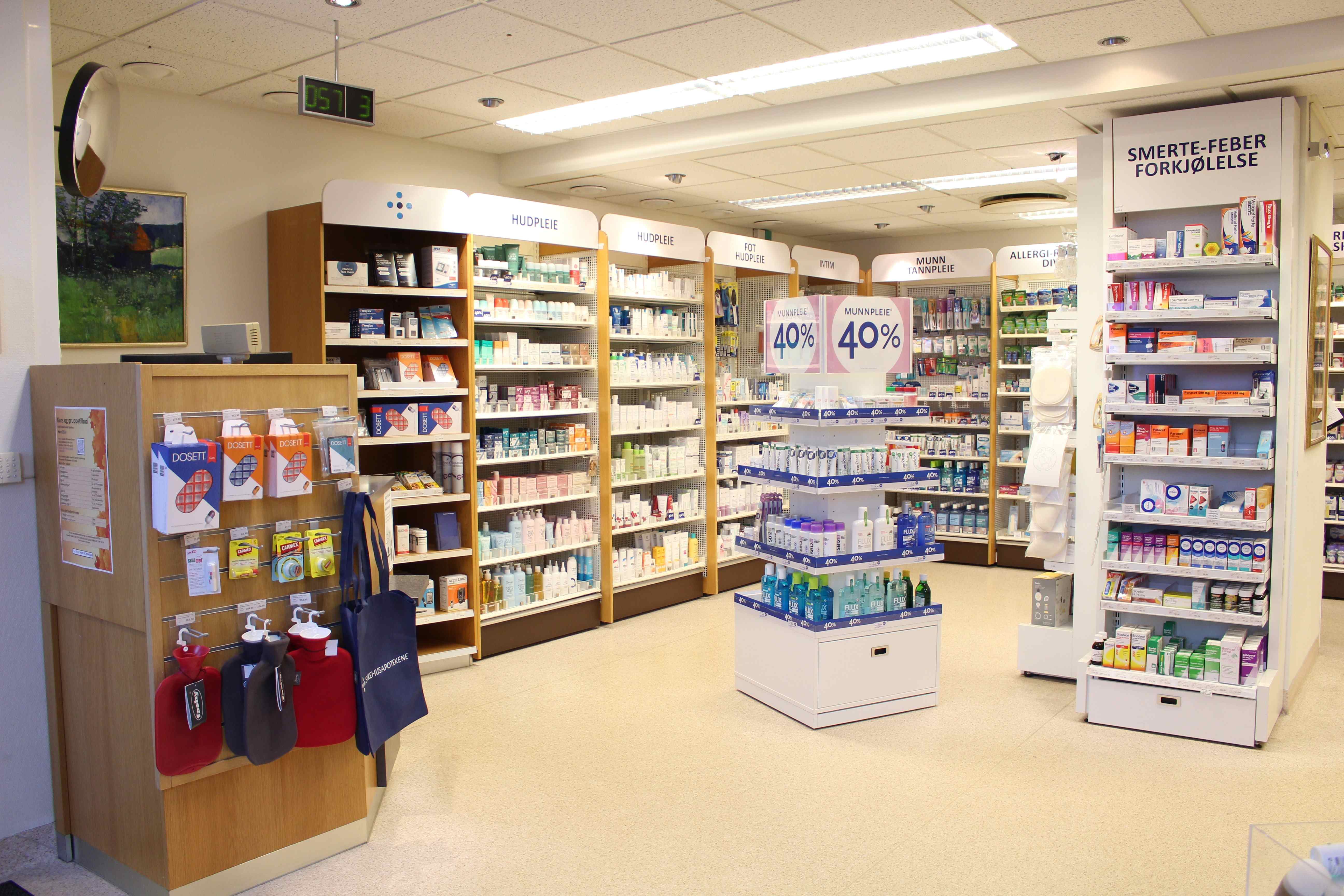 Lyst og oversiktlig apoteklokale med flere reoler fylt med apotekvarer og helseprodukter. I midten av lokalet står en utstillingshylle med produkter, og langs veggene er det hyller med blant annet hudpleieprodukter, kosttilskudd og reseptfrie legemidler. Ved inngangen til venstre er det et stativ med brosjyrer og handlenett. På bakveggen er det synlige kampanjeskilt med «40 %», og på høyre side henger et skilt med teksten «SMERTE/FEBER FORKJØLELSE». Lokalet har lyst gulv, god belysning og brede gangarealer.