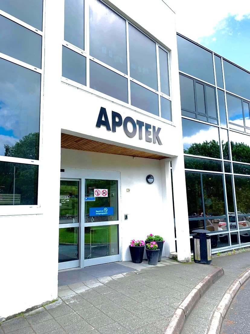 Inngangsparti til et apotek i en moderne bygning med store glassvinduer. Over inngangen er ordet «APOTEK» montert med store mørke bokstaver på en lys fasade. Vinduene reflekterer blå himmel og grønne trær utenfor. Bygningen har et rent og moderne uttrykk med glass-, tre- og betongelementer.