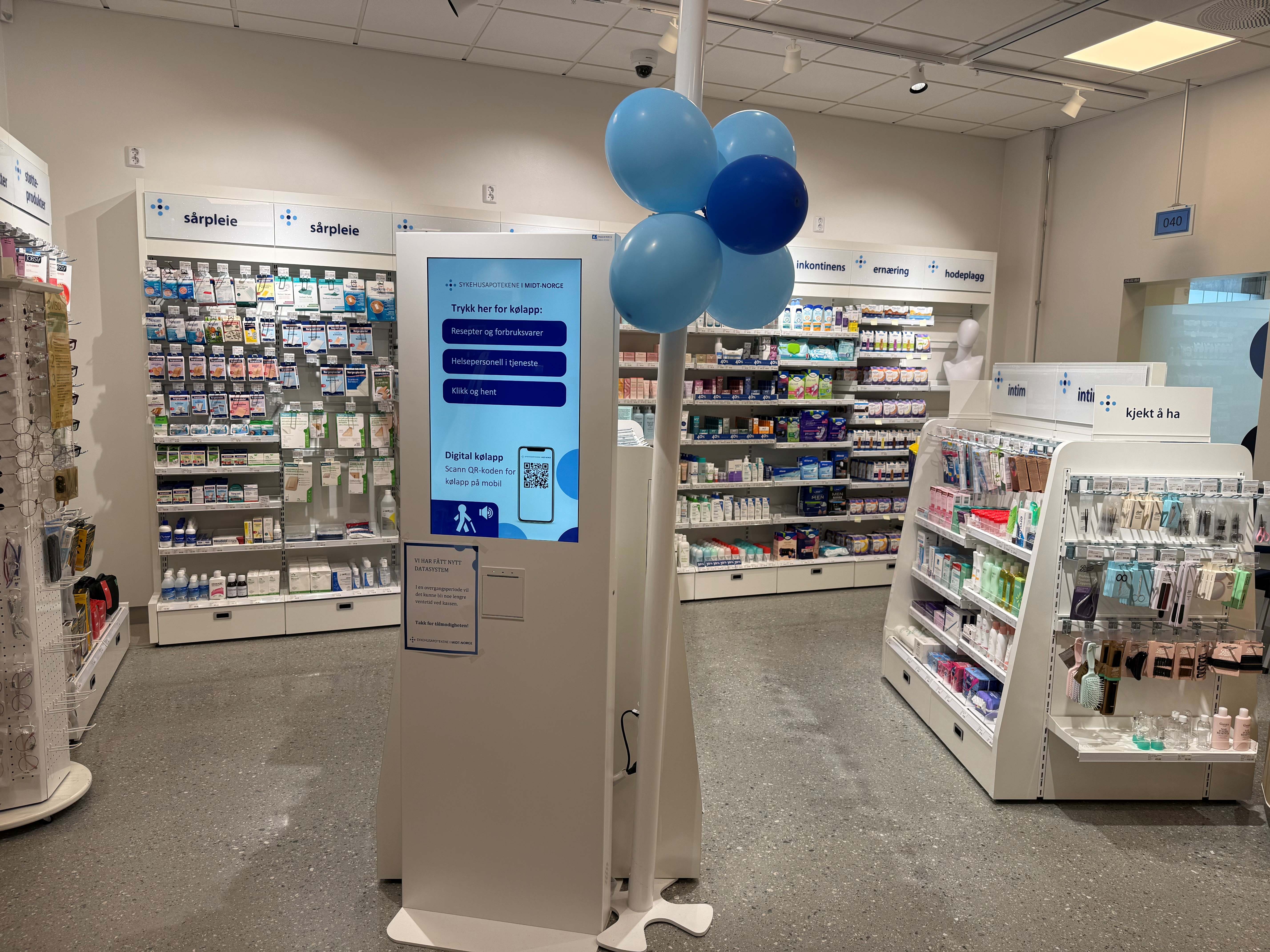 Interiør fra et apotek med varehyller langs veggene og produktutstillinger i forgrunnen. Midt i bildet står en digital informasjonsskjerm med valg for ulike tjenester og en QR-kode til digital kølapp. Rundt skjermen er det hyller med blant annet hudpleieprodukter, egenpleieartikler og apotekvarer. Til høyre står en produktreol med intimprodukter, og over skjermen henger blå ballonger. Lokalet er lyst, moderne og godt opplyst.