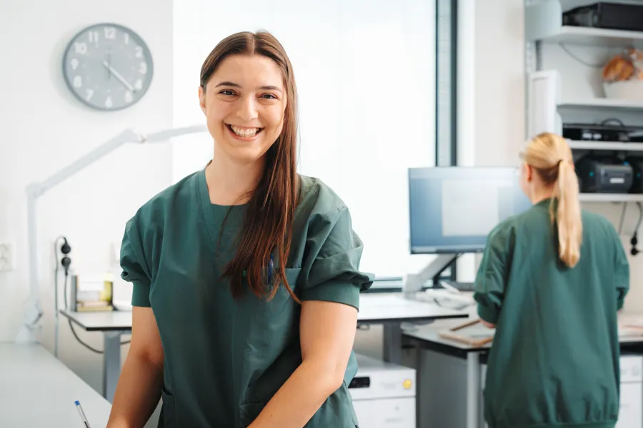 Person med langt hår og grønt arbeidstøy sitter i et lyst miljø. Bildet er beskåret i en sirkelform, og i bakgrunnen er det synlig en dataskjerm, skrivebord og deler av en annen person ved en arbeidsstasjon.